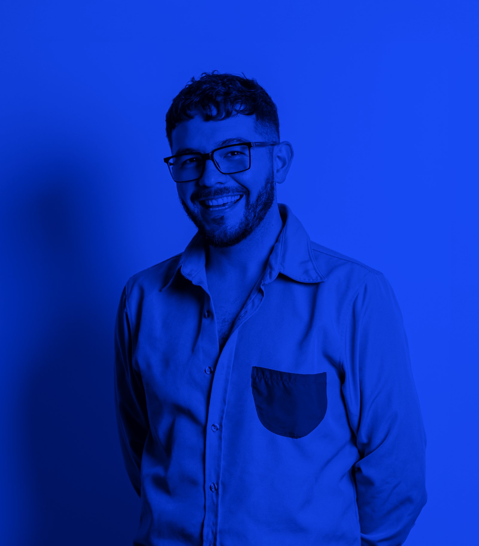 Juan sonriendo, con lentes y una camisa con bolsillo, en azul, contra una pared azul lisa