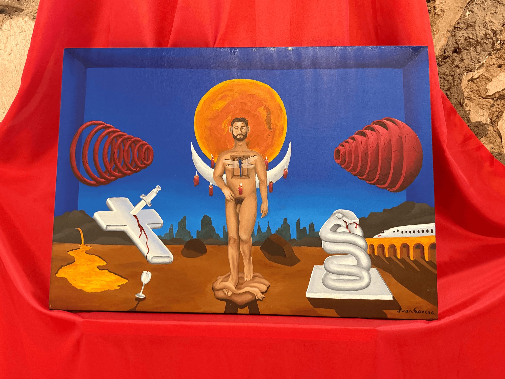 La pintura Génesis completa, apoyada sobre tela roja: un hombre con una libélula en el pecho, una luna creciente y un sol naranja detrás de él